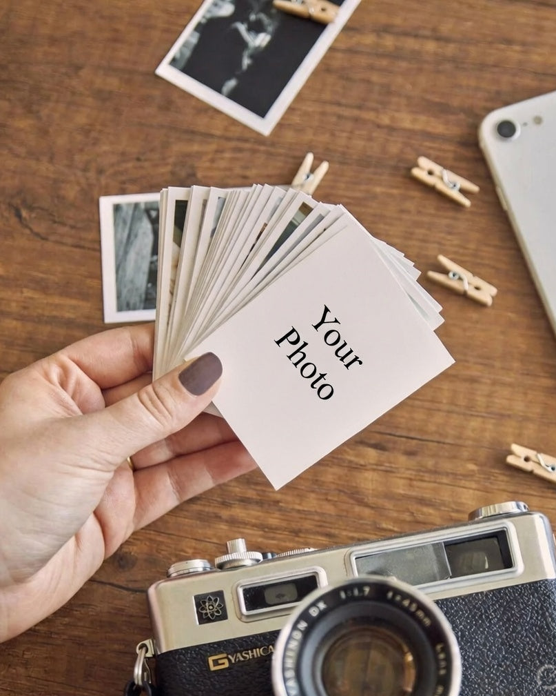 Hand holding photo cards with 'Your Photo' text, vintage camera, and smartphone on a wooden surface.