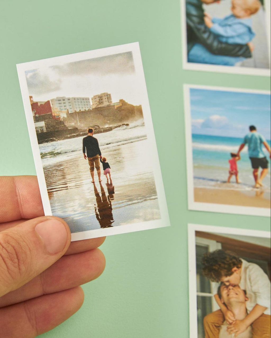 Hand holding a Polaroid photo of a man and child on a beach with more photos in the background.
