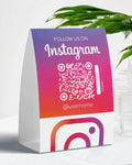 Colorful stand with Instagram QR code and username on a white surface with a plant in the background